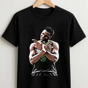 May include: Black t-shirt featuring a graphic of a basketball player with crossed arms and the number 5 in green. The word "TIMBERWOLVES" is visible on the player's wrist. The shirt is hanging on a wooden hanger.