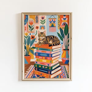 May include: A framed art print featuring a tabby cat resting atop a colorful stack of books. The illustration has a bohemian style, with potted plants, floral patterns, and a patterned rug. The color palette includes orange, blue, and green.