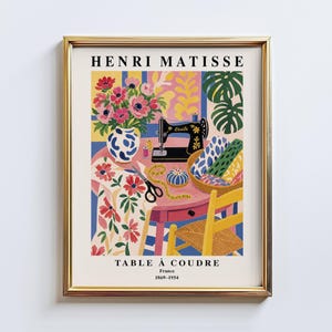 Peut inclure: Une affiche encadrée représentant une scène de couture colorée. L'œuvre d'art comprend une machine à coudre, des fleurs dans un vase, du tissu et le texte "HENRI MATISSE TABLE À COUDRE France 1869-1954". Les couleurs sont vives : rose, bleu, jaune et vert.