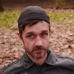 May include: A black cap with a brimless design. The cap is worn by a person with a beard and mustache, wearing a black leather jacket. The background features fallen leaves and a tree.