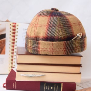 Puede incluir: Un gorro de cubo de lana a cuadros marrones, rojos y verdes con un alfiler plateado en el lateral. El gorro está encima de una pila de libros.