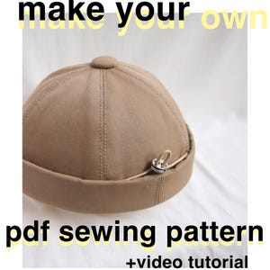 May include: A tan docker hat with a small metal loop detail. The text "make your own" is at the top, and "pdf sewing pattern + video tutorial" is at the bottom. The hat is made of a sturdy fabric.
