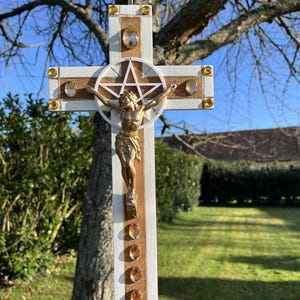 May include: A wooden cross featuring a white and natural wood finish, adorned with amber-colored faceted gems. A gold-toned figure is mounted at the center, positioned in front of a white circular frame containing a five-pointed star symbol.