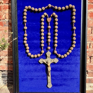 Peut inclure: Un chapelet encadré composé de perles en bois brun finement sculptées, disposées en rectangle sur un fond en velours bleu vif. Un crucifix métallique représentant le Christ est suspendu à la partie inférieure des perles.