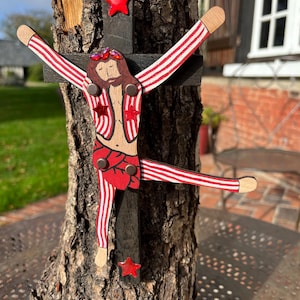 May include: A wooden crucifix featuring a dark-stained cross mounted on a rough tree trunk. A stylized figure with brown hair and red and white striped limbs is attached to the cross, accented with red star-shaped embellishments and red gems.