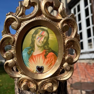 Peut inclure: Un cadre ovale de couleur dorée avec des détails de volutes ornementales entoure une impression d'une figure aux longs cheveux bruns, vêtue d'une robe bleue et rouge, avec un éclair rouge et bleu sur un œil et un petit emblème de cœur rouge.