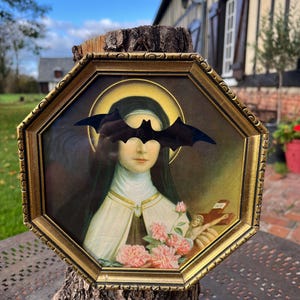 Peut inclure: Un cadre octogonal doré contenant un portrait religieux d'une figure en habit avec une auréole dorée. Une chauve-souris noire découpée est placée sur les yeux. La figure tient un petit crucifix en bois et un bouquet de roses roses dans ses mains.