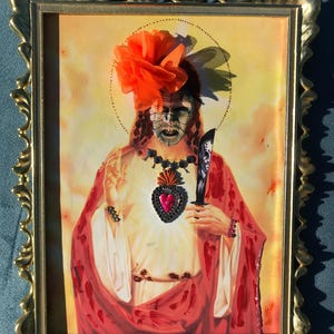 May include: A framed mixed media artwork featuring a figure with a grid-patterned face, wearing a bright orange fabric flower, a beaded necklace with a heart pendant, and holding a knife, set against a yellow background in a gold frame.