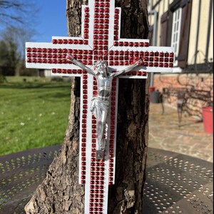 Peut inclure: Une croix en bois blanc ornée de rangées de petits strass carrés rouges, avec une figurine en métal argenté représentant Jésus-Christ fixée au centre, le tout monté verticalement sur un morceau d'écorce d'arbre brun à la texture brute.