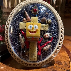Peut inclure: Une plaque murale ovale avec un fond en mosaïque bleue et une bordure en treillis crème. Une croix faite d'éponges jaunes est centrée, ornée d'une figurine de Bob l'éponge, de divers coquillages, de corail rouge et d'un petit emblème en cœur.