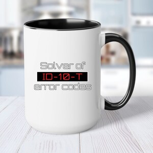 May include: White ceramic mug with a black rim and handle. The mug features a black and white graphic that says "Solver of ID-10-T error codes".