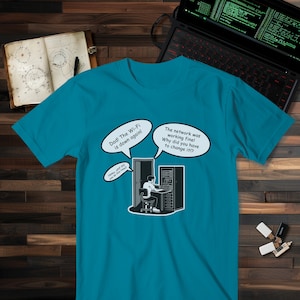 Può includere: T-shirt turchese con un fumetto umoristico sui problemi del Wi-Fi. Il design presenta fumetti con frasi come "Papà! Il Wi-Fi è di nuovo giù!" e "La rete funzionava bene!"