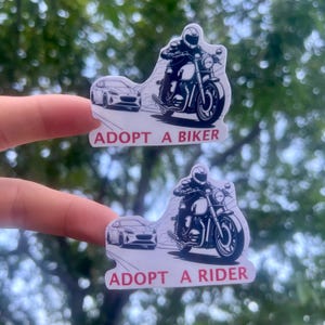 May include: Two white stickers with black and white illustrations of a motorcycle rider and a car. The top sticker says "Adopt a Biker" and the bottom sticker says "Adopt a Rider".