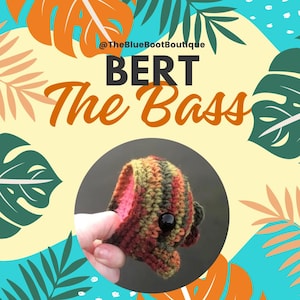 May include: A crocheted bass fish toy named "BERT The Bass" in shades of green, brown, orange, and pink. The toy has a large black button eye. The image has a tropical background with palm leaves.