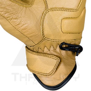 Peut inclure: Gros plan sur un gant en cuir beige avec des d&eacute;tails noirs. Le gant pr&eacute;sente des coutures, un clip noir et une sangle Velcro. Le bord inf&eacute;rieur est bord&eacute; de noir. Le mot "THE" est partiellement visible.