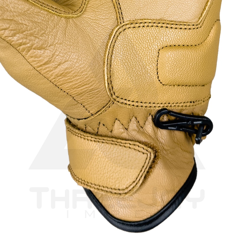 Peut inclure: Gros plan sur un gant en cuir beige avec des d&eacute;tails noirs. Le gant pr&eacute;sente des coutures, un clip noir et une sangle Velcro. Le bord inf&eacute;rieur est bord&eacute; de noir. Le mot "THE" est partiellement visible.