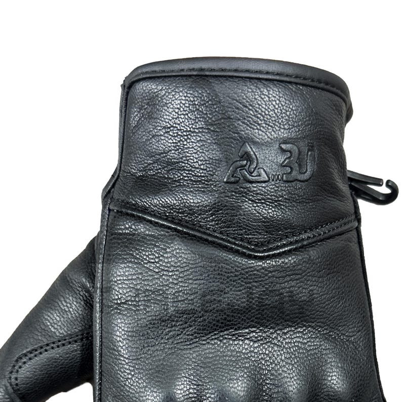 Peut inclure: Gros plan d'un gant en cuir noir. Le gant pr&eacute;sente une surface textur&eacute;e et une couture. La partie sup&eacute;rieure du gant comporte un logo en relief. Un clip en plastique noir est attach&eacute; sur le c&ocirc;t&eacute;.