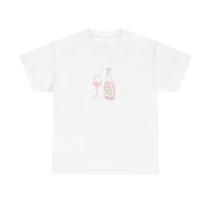 Puede incluir: Camiseta blanca con una ilustración en acuarela de una botella de vino y una copa de vino, ambas llenas de líquido rosa. La botella tiene una etiqueta con texto y una pequeña ilustración. El diseño está centrado en la camiseta.