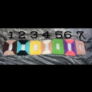 May include: Seven colourful, fluffy wristbands are displayed against a dark background. Each wristband has a rectangular opening in the centre. The wristbands are in shades of brown, teal, yellow, peach, green, purple, and pink.