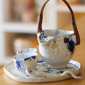 Könnte beinhalten: Ein weißes Keramik-Tee-Set mit blauen und braunen Akzenten. Das Set beinhaltet eine Teekanne mit Ledergriff, zwei Tassen und ein Tablett. Die Teekanne und die Tassen haben eine gesprenkelte Glasur.