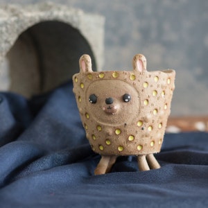 May include: A brown ceramic planter shaped like a bear with a textured surface and yellow polka dots. The planter has a bear face with black eyes, a black nose, and small pink ears. It stands on three small legs.