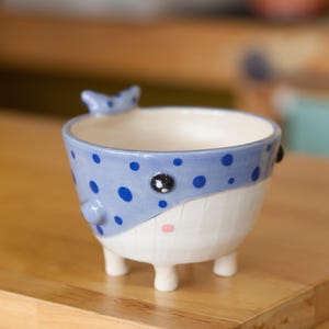 Puede incluir: Un pequeño macetero de cerámica azul y blanco con forma de ballena con ojos negros y manchas azules. El macetero tiene cuatro pequeñas patas y una abertura redonda ancha.