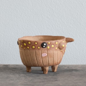 May include: A small, brown ceramic planter with three legs and a yellow and brown polka dot design. The planter has a black button nose and a pink mouth.