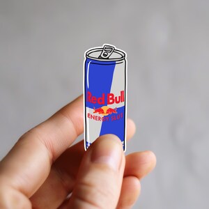 Op de afbeelding: Een sticker van een Red Bull energiedrankblikje met een blauw en zilver ontwerp. De tekst "Red Bull" is rood en de tekst "ENERGY SLUT" staat eronder. Het blikje wordt tussen de vingers gehouden.