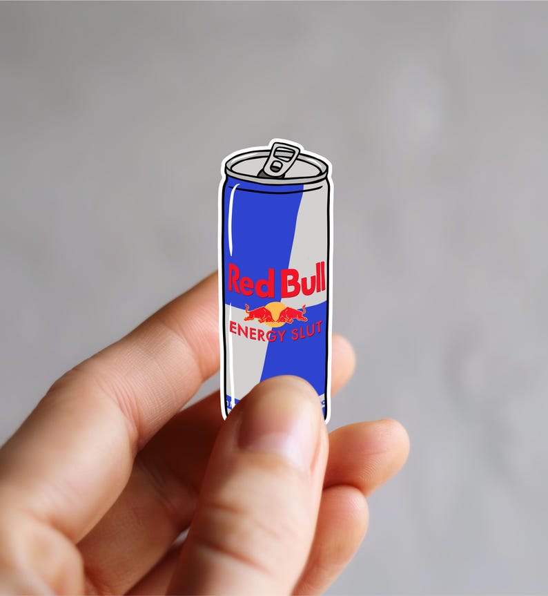 Op de afbeelding: Een sticker van een Red Bull energiedrankblikje met een blauw en zilver ontwerp. De tekst "Red Bull" is rood en de tekst "ENERGY SLUT" staat eronder. Het blikje wordt tussen de vingers gehouden.
