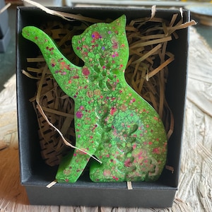 May include: A cat-shaped resin ornament in a vibrant lime green color, featuring embedded pink and iridescent glitter particles. The ornament is displayed inside a black cardboard gift box filled with brown paper shred filler material.