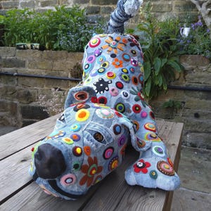 May include: A whimsical, handmade dog sculpture crafted from denim fabric. The dog is adorned with colorful felt flowers, buttons, and circular embellishments. It has a black nose and a striped tail, resting on a wooden surface.