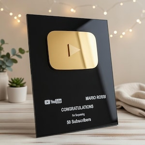 May include: A black acrylic YouTube play button award with a gold play button and white text. The text reads "Congratulations For Surpassing 50 Subscribers" and the name "Mario Rossi". The award is on a wooden surface, with fairy lights in the background.