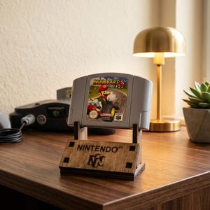 Pode incluir: Um cartucho de videogame Mario Kart 64 cinza para o console Nintendo 64, exibido na vertical em um suporte de madeira marrom escuro. O suporte apresenta o texto gravado NINTENDO 64 e o logotipo N64 no painel frontal da base.