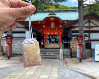 Puede incluir: Un pequeño amuleto omamori de tela rectangular con un degradado de amarillo pálido a rosa suave. Está decorado con grullas bordadas en oro y texto en japonés, rematado con un cordón blanco atado en un nudo decorativo en la parte superior.