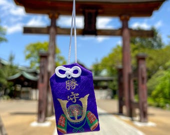 Puede incluir: Un amuleto omamori de tela morada con un cordón blanco en la parte superior y caracteres japoneses bordados en oro sobre un gráfico de casco estilizado con detalles en rojo, verde y dorado. El amuleto rectangular tiene un nudo decorativo blanco.