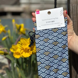 May include: A blue drawstring bag with a white and gold wave pattern. The bag has a square shape and is made of fabric. The text on the bag reads "Square Drawstring Bag" and "正方形巾着袋".