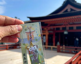 Puede incluir: Un amuleto japonés omamori con una ilustración de ciervo, una campana y texto en japonés. El amuleto está en una funda de plástico transparente, frente a un edificio tradicional japonés con un puente rojo y cielo azul.