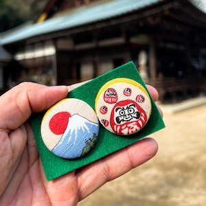 以下が含まれることがあります： 緑色のフェルト地の背景に、布製のピンバッジが2つ。1つのバッジは、赤と白の富士山が描かれている。もう1つのバッジは、赤と白の伝統的な日本のだるま人形が描かれている。