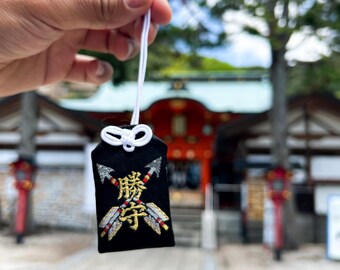 Puede incluir: Un amuleto omamori de tela negra con dos flechas cruzadas bordadas en hilo plateado, rojo y blanco, con los caracteres kanji japoneses dorados para la victoria, suspendido por un cordón blanco con un nudo decorativo en la parte superior.