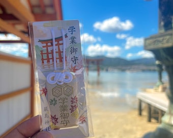 Puede incluir: Un amuleto omamori japonés en una funda de plástico transparente. El amuleto tiene detalles dorados y rojos con texto japonés. El fondo muestra una masa de agua, una puerta torii y un cielo azul.