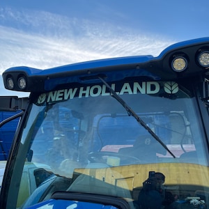 Puede incluir: Una cabina de tractor azul con un parabrisas y un logotipo de New Holland. El logotipo es blanco con un diseño de hoja verde.