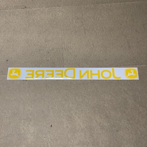 May include: A white rectangular sticker featuring the yellow text "JOHN DEERE" in a bold, sans-serif font. The sticker has a yellow deer logo on each end. The sticker is designed for application on a vehicle or equipment.