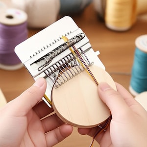 Könnte beinhalten: Ein weißes und silbernes Webwerkzeug mit Holzsockel wird von Händen gehalten. Das Werkzeug hat Metallhaken und wird mit bunten Fäden verwendet. Garnrollen in Lila, Gelb und Türkis sind im Hintergrund.