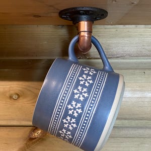 May include: A blue ceramic mug with a white snowflake design suspended from a black pipe fitting. The mug hangs from a copper pipe attached to a wooden beam. The mug has a cream-coloured base.