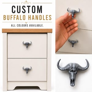 May include: A set of grey buffalo skull-shaped drawer handles installed on a two-drawer nightstand. The text CUSTOM BUFFALO HANDLES ALL COLOURS AVAILABLE is displayed above. The handles feature a detailed animal skull design with curved horns.