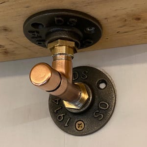 Könnte beinhalten: Ein Kupfer- und Messingrohranschluss, montiert auf einer Holzoberfläche. Der Anschluss ist an einem schwarzen Metallflansch mit dem Text "DS 1914 OS" befestigt. Das Design deutet auf eine industrielle oder rustikale Ästhetik hin.