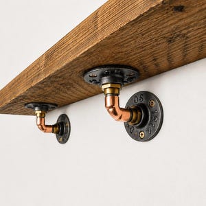 Könnte beinhalten: Ein Holzregal, das von zwei Halterungen im Industriestil getragen wird. Jede Halterung besteht aus einem schwarzen, runden Metallflansch mit der Aufschrift 1014 und OS, der mit einem kupferfarbenen Winkelrohrstück an der Unterseite des Holzes befestigt ist.