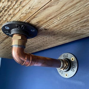 May include: A copper pipe shelf bracket featuring two black circular metal flanges with the letters DS and 19 stamped on them. The bracket connects a wooden shelf to a wall, secured with silver and gold-colored screws at each mounting point.