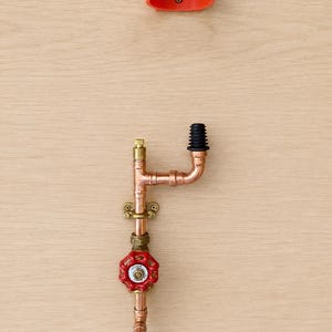 Op de afbeelding: Een koperen leidingconstructie met een rode ronde ventielknop, een messing montagebeugel en een zwarte geribbelde rubberen dop. Boven de leidingconstructie bevindt zich een losse, U-vormige oranje kunststof montageclip met een schroef.