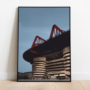 Könnte beinhalten: Gerahmter Kunstdruck mit einem modernen Stadion. Das Bild zeigt ein Stadion mit schwarzem Dach und roten Stützen vor blauem Himmel. Das Gebäude hat ein einzigartiges zylindrisches Design, mit Personen am unteren Rand.